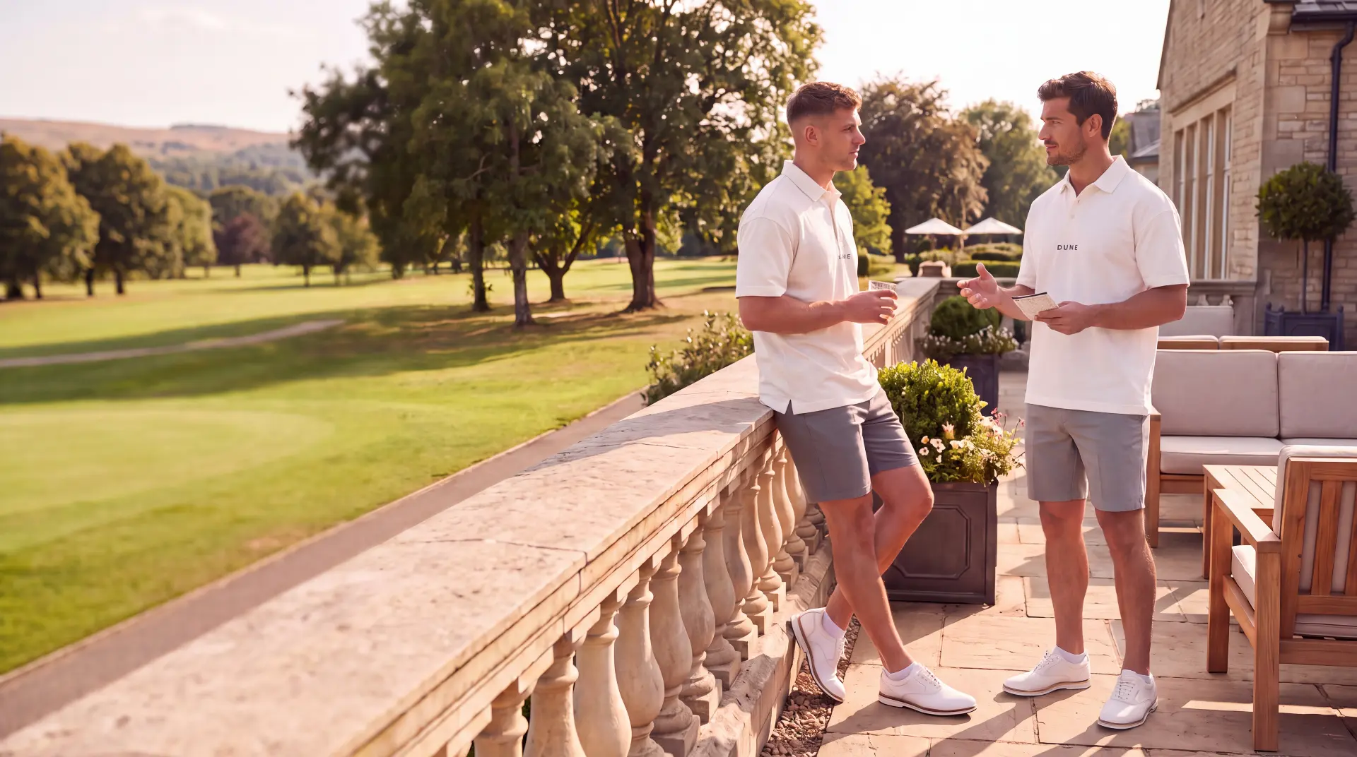Two golfers in white DUNE polos talking on a stone clubhouse terrace overlooking a Dubai golf course